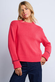 A female model wearing a pink oversized sweater with rolled rib cuffs and contrast stitching, paired with dark trousers.