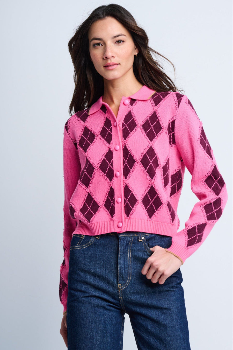 A woman wearing a pink argyle intarsia cardigan with a collared neckline and six-button front, paired with blue jeans.