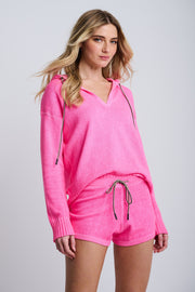A female model wearing a neon pink knit hoodie and shorts set with ribbed finishing at the bottom hem and contrast pulls at the waistband.