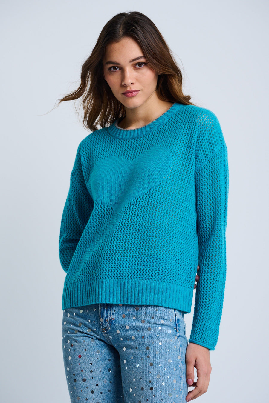 A woman wearing a blue cashmere sweater with an open stitch and heart intarsia at the center front, contrast tipping at the neckline and rolled cuffs, paired with dotted light blue jeans.