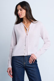 A woman wearing a light pink, slim fit cashmere sweater with an open V-neck, heart-shaped buttons, and ribbed edges at the cuffs and bottom hem.