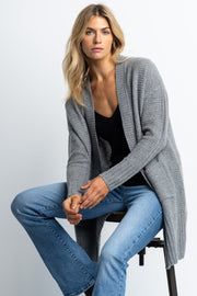 A female model wearing a gray open-front cashmere cardigan with waffle-stitch texture and ribbed finishing, paired with a black top and blue jeans.