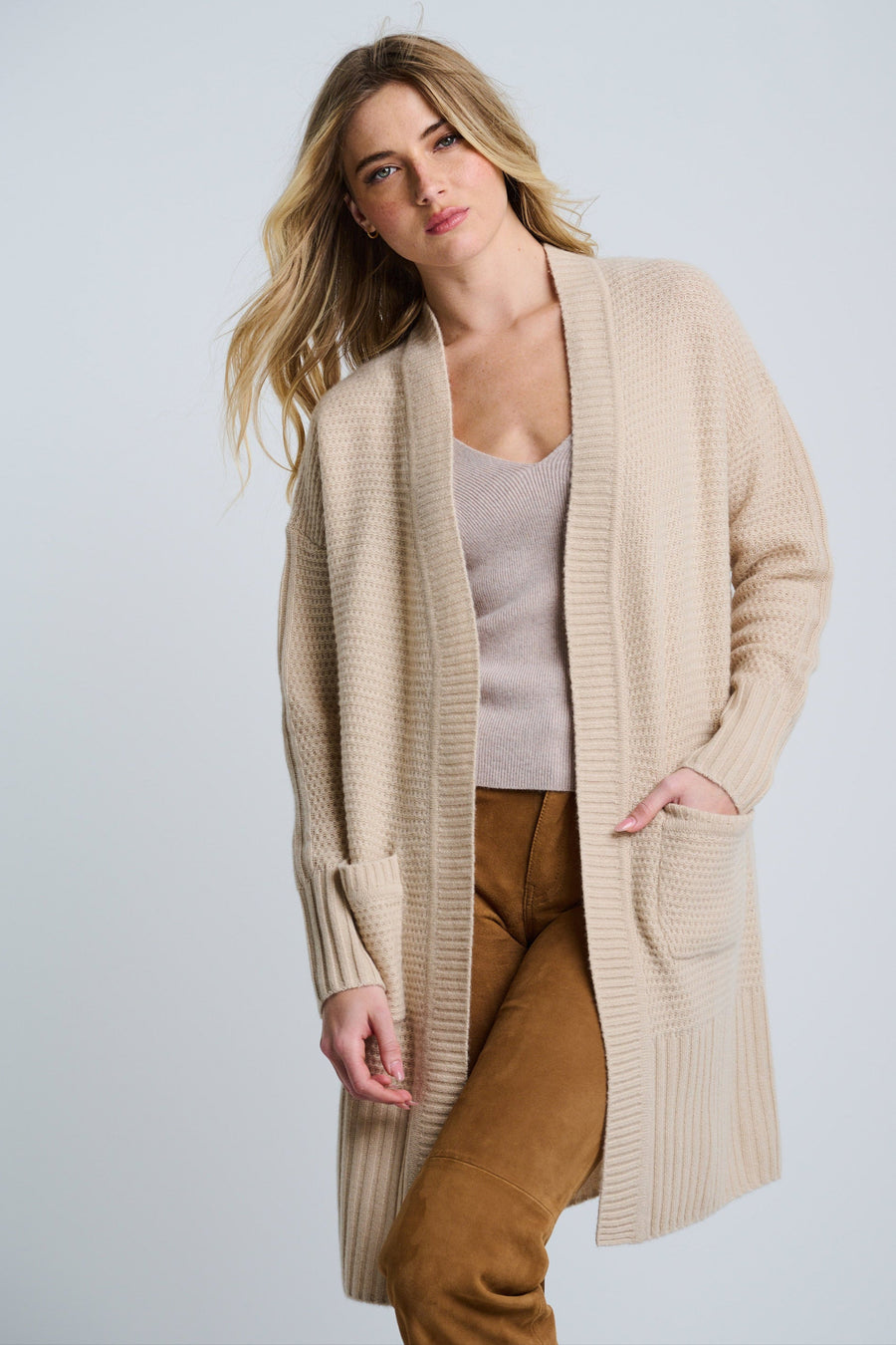 A female model wearing a beige open-front long cardigan with patch pockets, over a white top, paired with brown trousers.