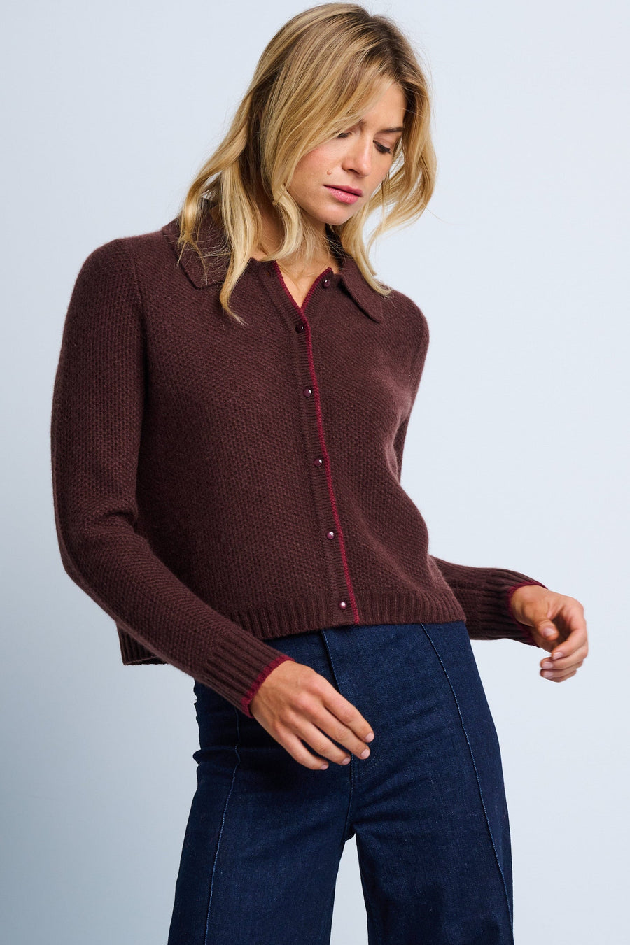 A woman wearing a brown cashmere cardigan with a tailored collar, contrast tipping, and a relaxed drape. She is also wearing blue jeans.