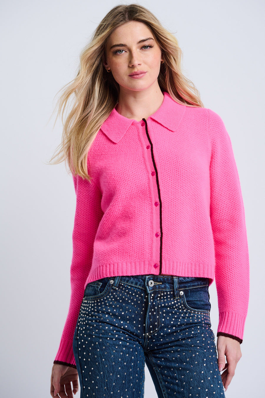 A female model wearing a pink cashmere cardigan with a tailored collar, contrast tipping, and a relaxed drape. She is also wearing blue jeans with a dotted pattern.