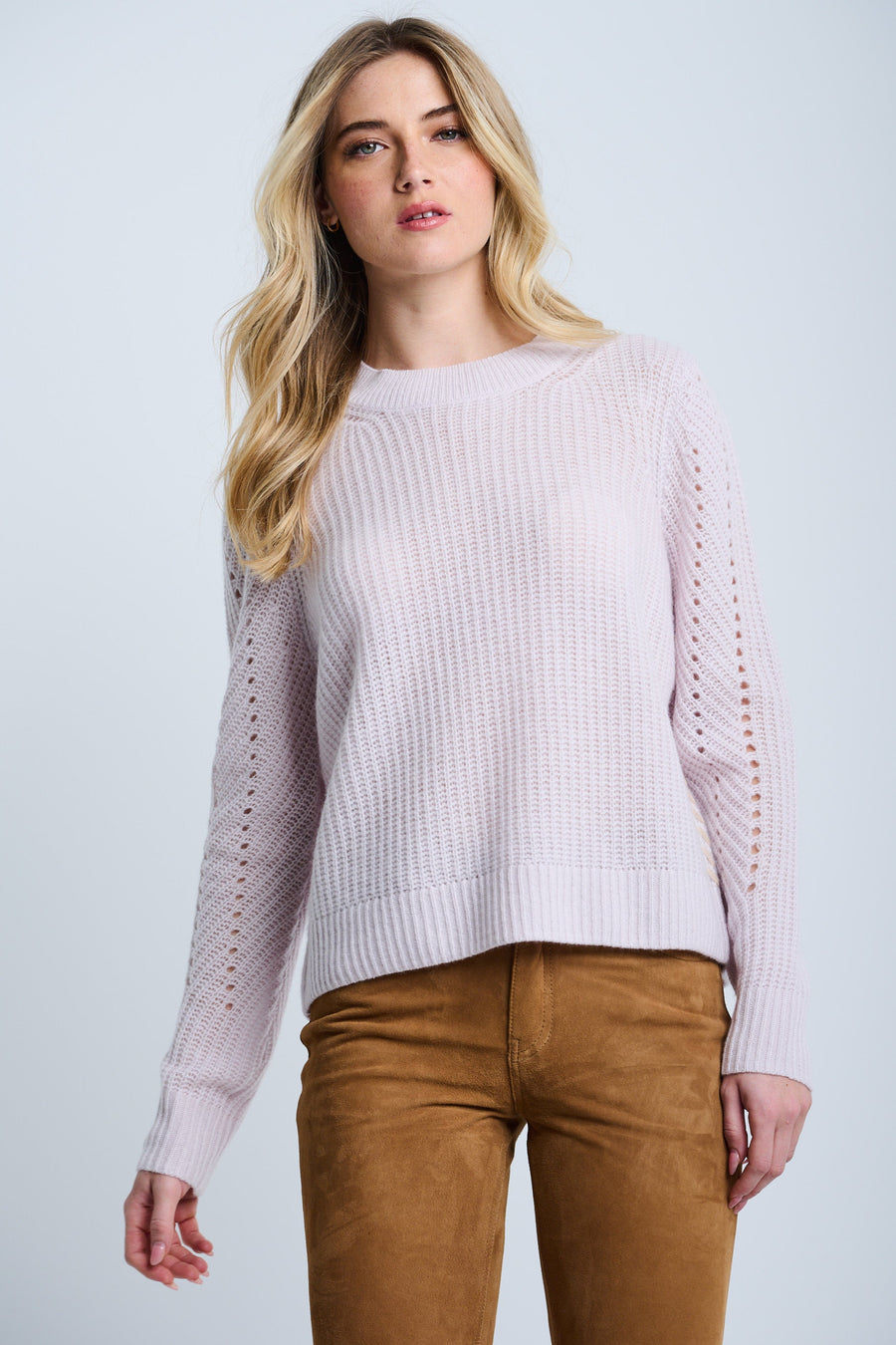 A woman wearing a pink Luxe Softie crewneck sweater with ribbed edges and pointelle detail on the sleeves, paired with brown pants.