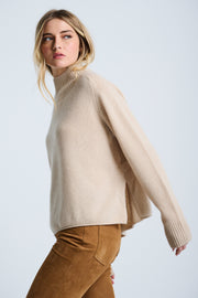 A woman wearing a beige oversized turtleneck sweater with side slits and colorblock cuff accents, paired with brown pants.