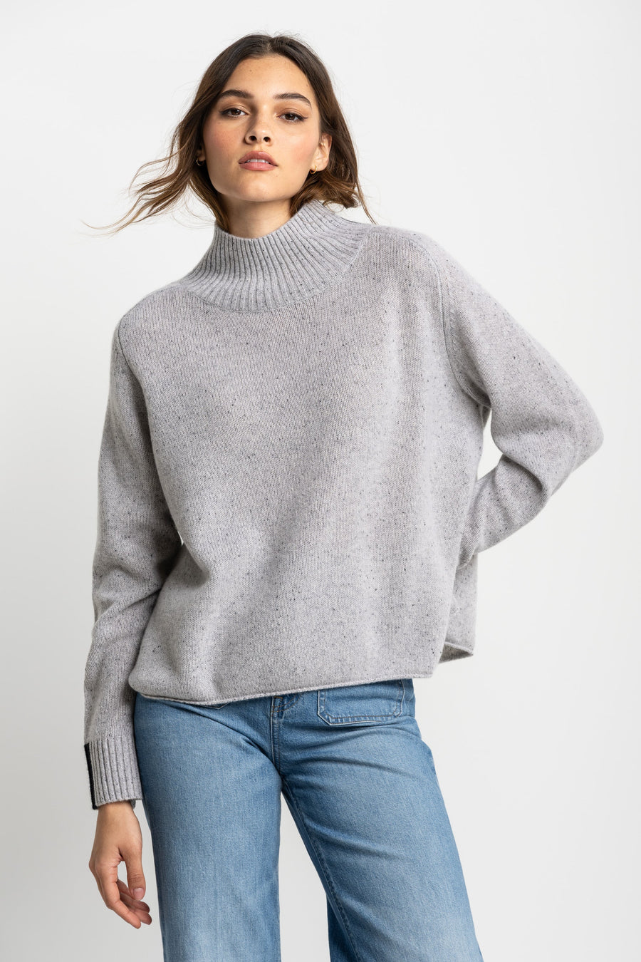 A woman wearing a grey oversized pullover with a high ribbed neckline and colorblock cuff detailing, paired with blue jeans.