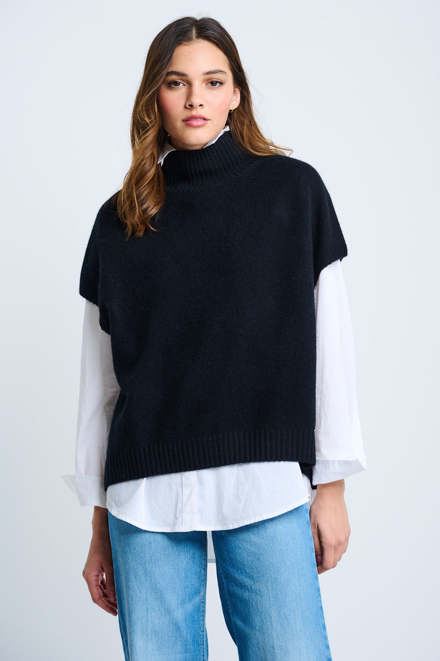 A woman wearing a black high rib neck, short sleeve pullover with drop shoulder and ribbed finishing at the bottom hem, paired with a white shirt and blue jeans.