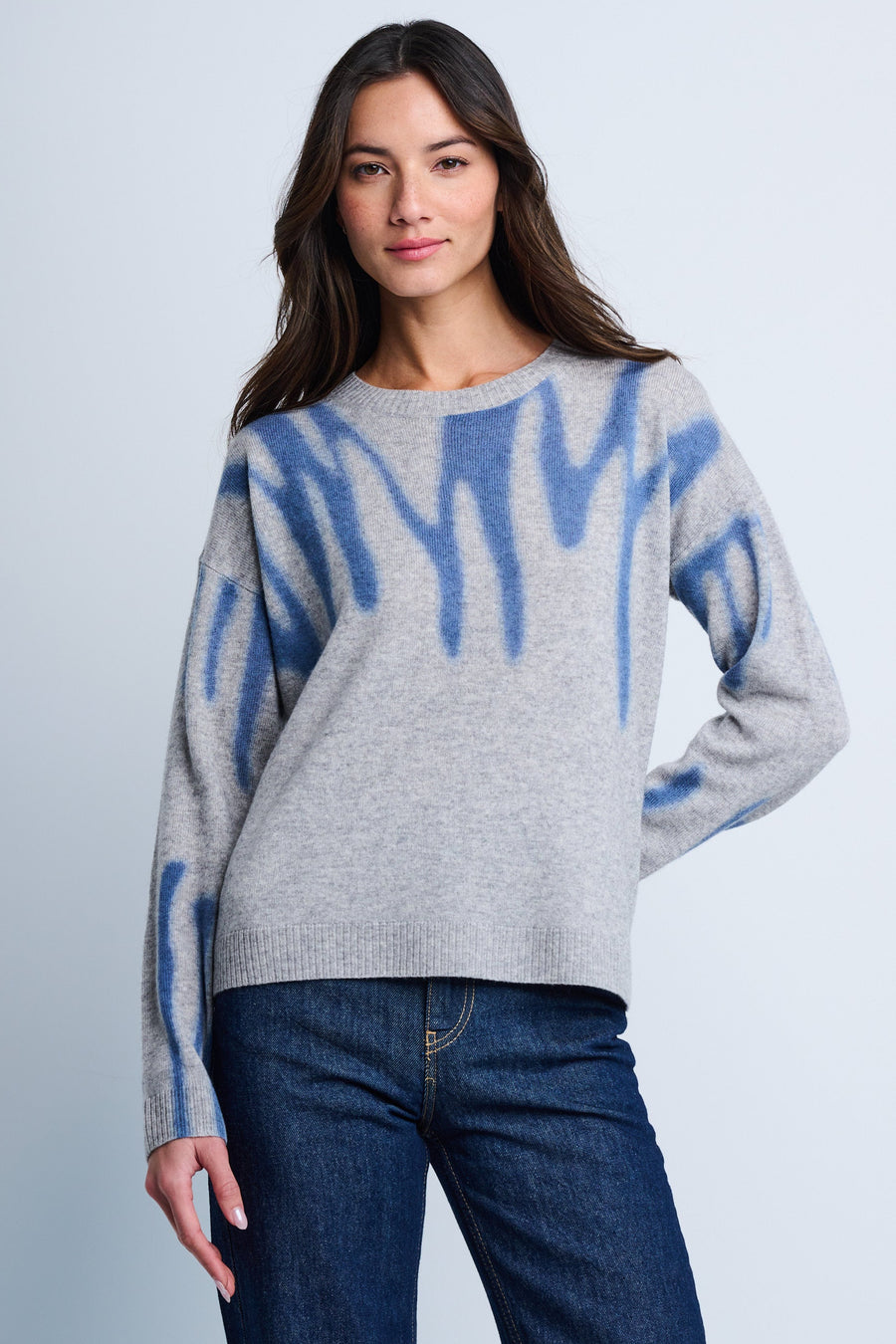 A female model wearing a relaxed fit, crew neck sweater with an all-over mod print in blue and gray tones, with drop shoulder sleeves and ribbed cuffs and bottom hem, paired with blue jeans.
