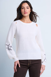 A woman modeling a white crew neck sweater with rib-stitch texture, shoulder splice detail, and contrast hand-dash embroidery on the sleeves.