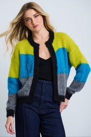 A female model wearing a colorblock striped open cardigan in blue, green, and gray tones, paired with a black top and blue jeans.