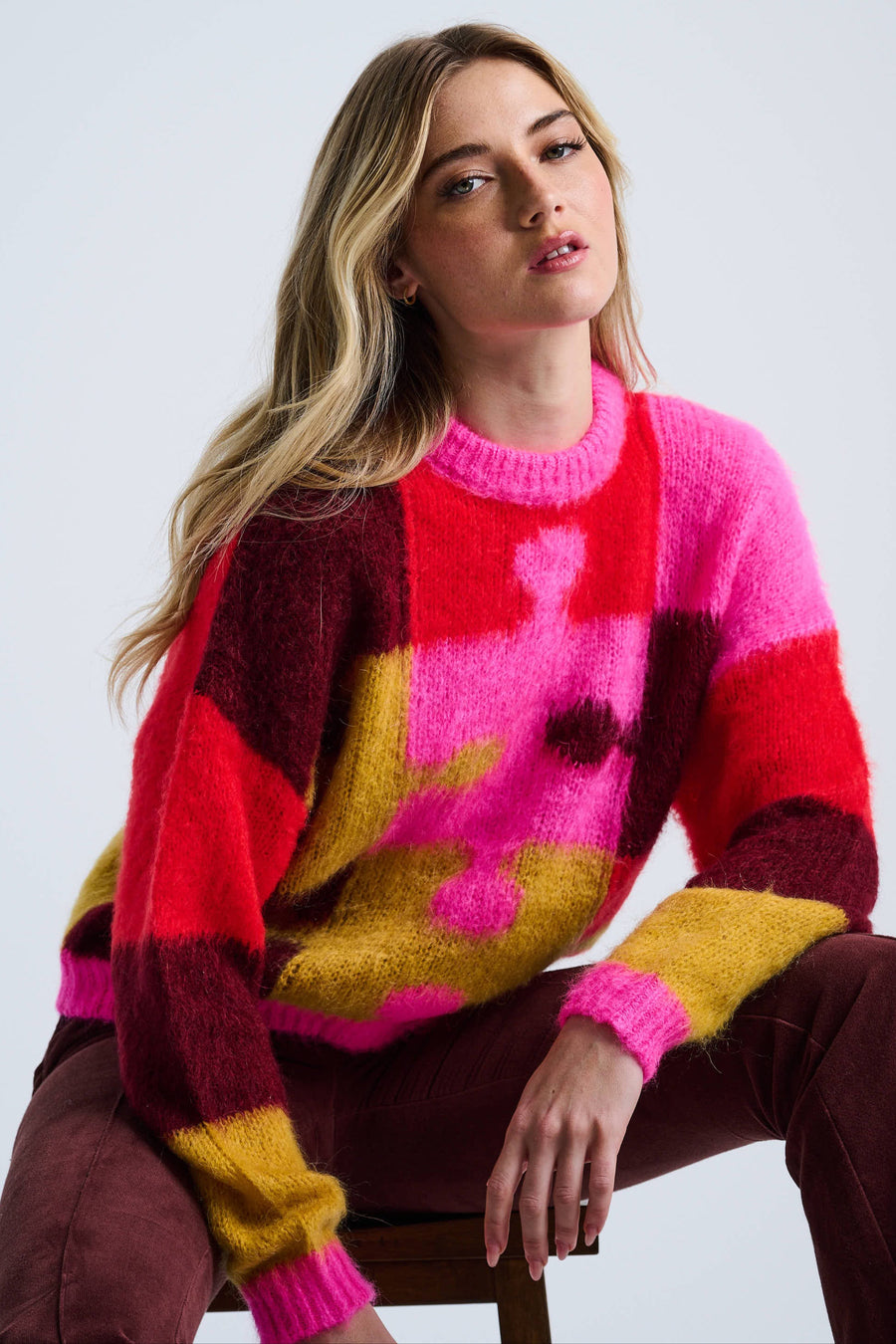 A woman sitting and modeling a colorful intarsia pullover sweater with a relaxed crewneck silhouette, predominantly in red and yellow tones.