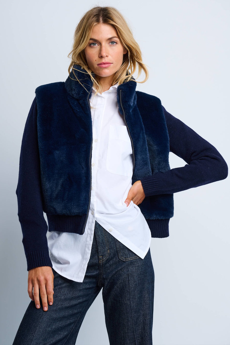 A female model wearing a navy blue zip-up jacket with a high collar and ribbed cuffs, paired with a white shirt and blue jeans.