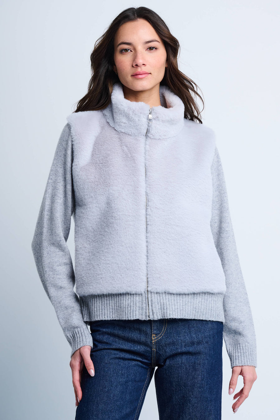 A female model wearing a light gray zip-up faux fur jacket with a high collar, ribbed cuffs, and pockets, paired with blue jeans.