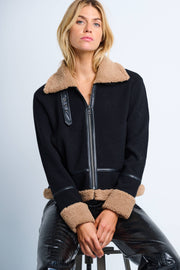 A woman sitting on a stool wearing a black zip-up jacket with faux fur at the collar, hem, and cuffs, along with faux leather trim and pockets.