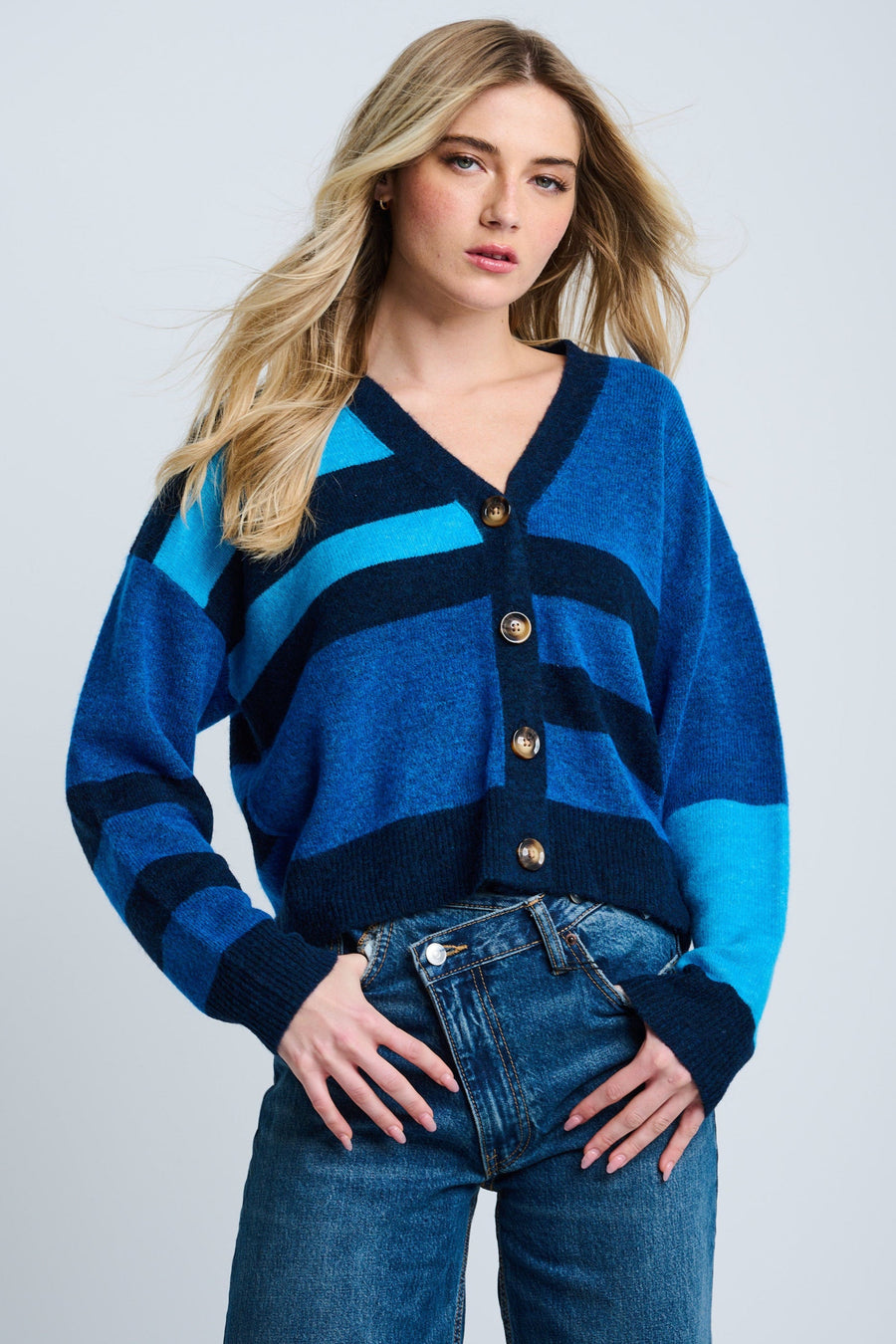 A female model wearing an oversized, colorblocked blue cardigan with a round neckline and four-button closure, paired with jeans.