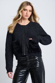 A female model wearing a black ribbed stitch jacket with plush faux fur pockets and a zip-up silhouette, paired with black leather pants.