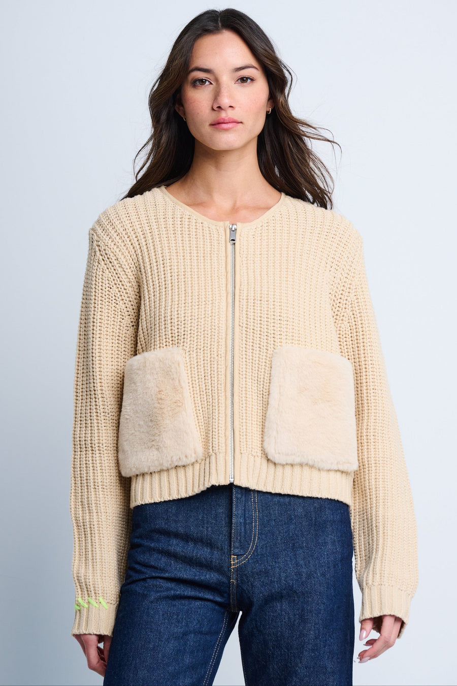 A female model wearing a beige zip-up jacket with plush faux fur pockets and ribbed stitch details, paired with blue jeans.