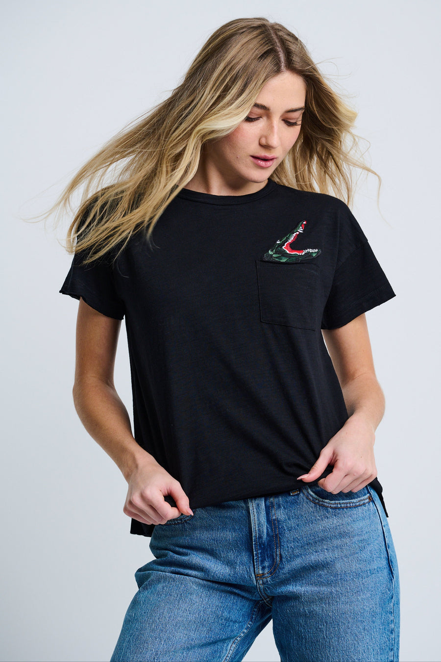A female model wearing a black short-sleeve crew t-shirt with an embroidered alligator applique above the chest pocket, paired with blue jeans.