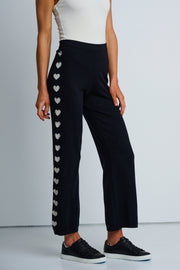 A model wearing black track pants with heart intarsia down the leg, paired with a white top and black sneakers.