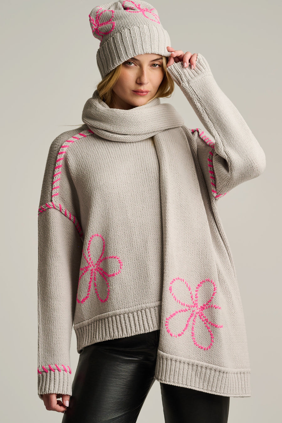 A woman models a chunky knit sweater with a matching knit beanie. The sweater is a light oatmeal beige color with bright pink embroidered flower motifs and decorative stitching along the shoulders and sleeves.