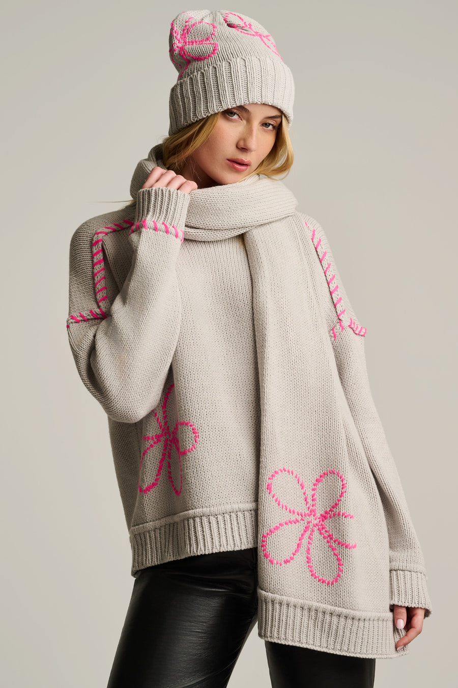 A woman models a chunky knit winter set featuring a light beige sweater with bright pink floral and geometric patterns, a matching beige scarf, and a beige beanie with pink details, set against a solid light gray background.