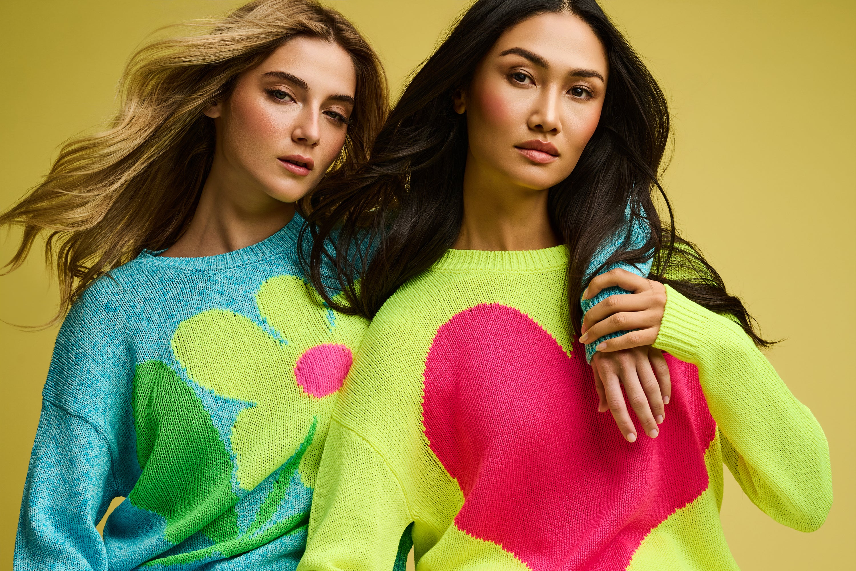 Two women wearing colorful sweaters with floral and heart designs on a yellow background