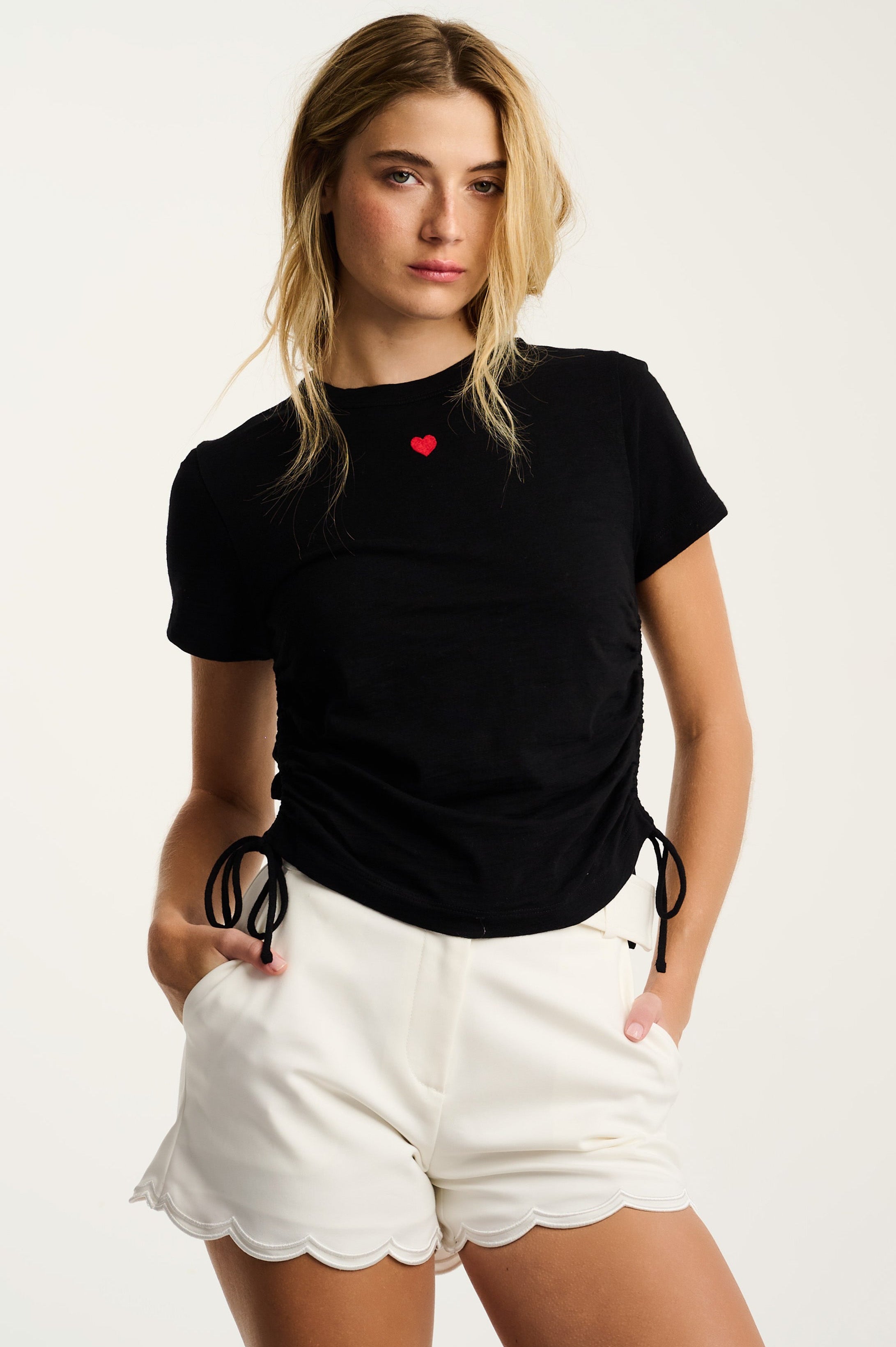 Woman wearing a black t-shirt with a red heart and white shorts on a plain background